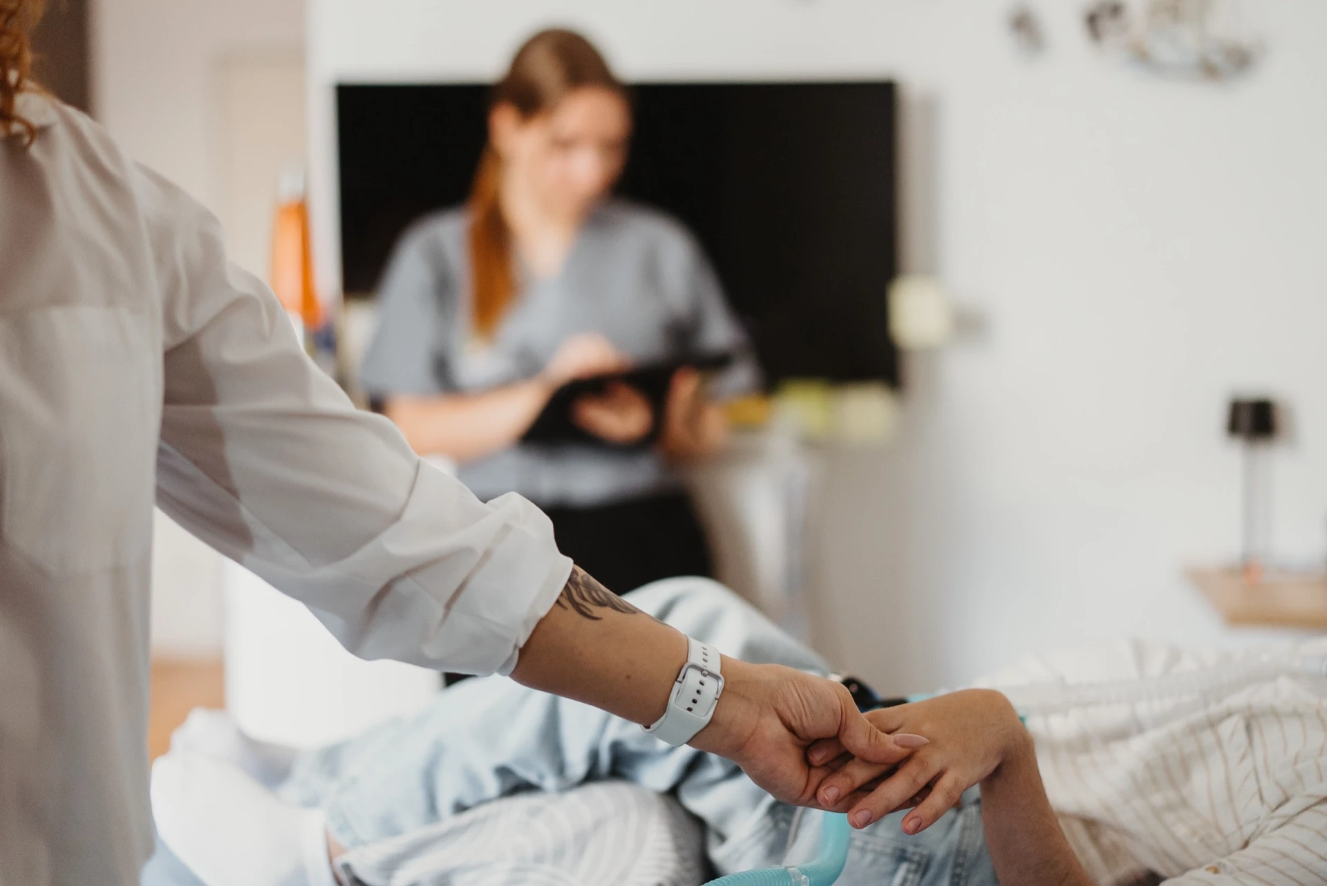 Eine Pflegekraft von Bernstein steht am Pflegebett einer Patientin und hält ihre Hand, am unteren Bildrand ein Beatmungsschlauch. Im unscharfen Hintergrund eine zweite Pflegekraft mit Tablet in einem Wohnraum.